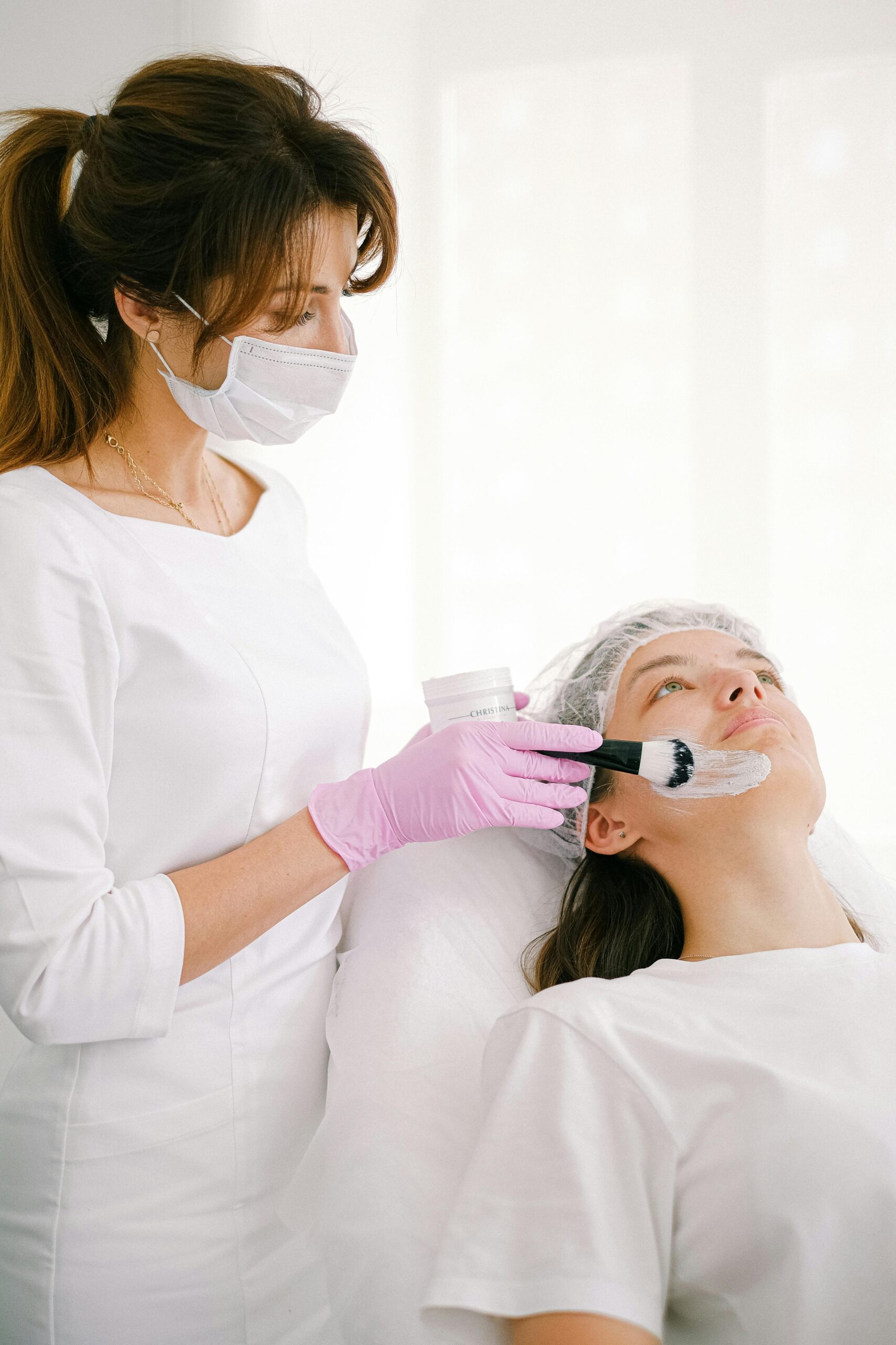 A beautician applies a facial mask to a customer in a bright spa room.
